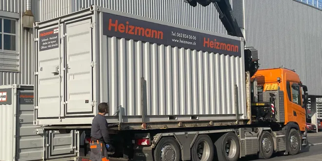 Chargement d'un conteneur marqué Heizmann par un camion avec grue pendant une journée ensoleillée