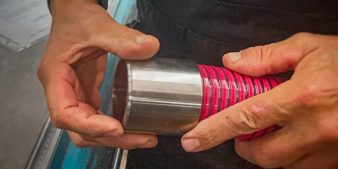 Person hält einen metallischen Zylinder mit einem roten flexiblen Schlauch in den Händen, im Hintergrund teilweise sichtbare Dokumente